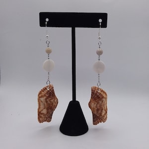 May include: A pair of dangle earrings featuring a brown seashell, a white round bead, and a smaller beige bead. The earrings are hanging from silver hooks.
