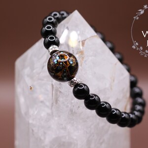 May include: Black beaded bracelet with a large, round, iridescent opal-like bead in the centre. The bracelet is displayed on a white crystal.