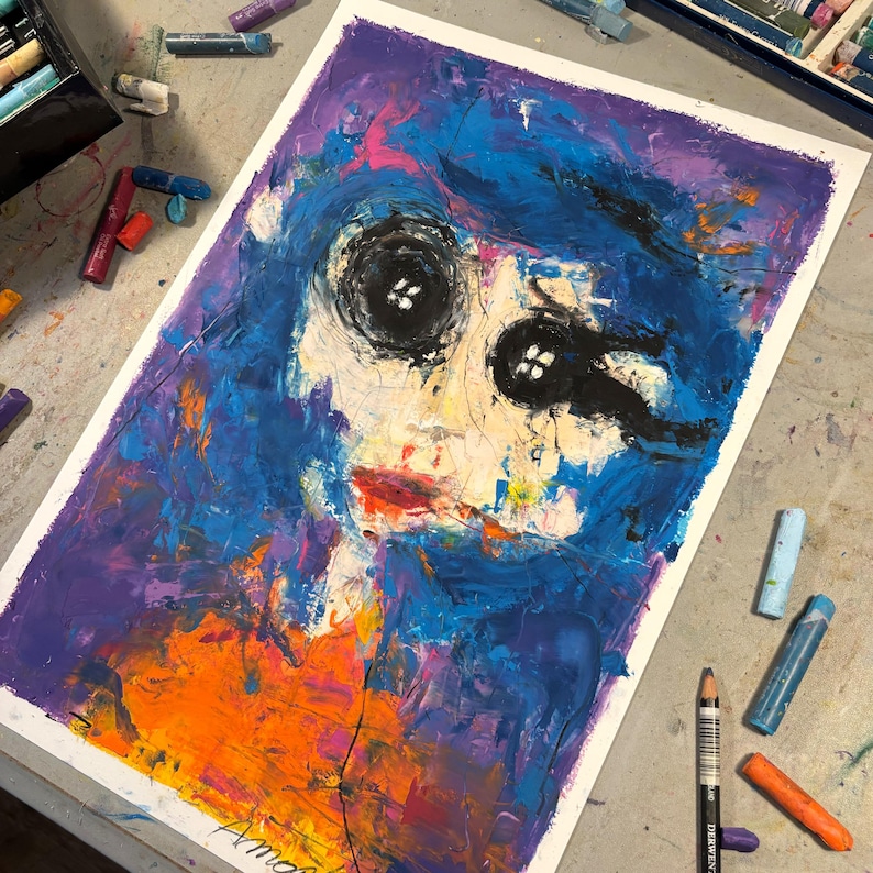 May include: Abstract painting of a face with button eyes, blue hair, and red lips. The artwork uses thick brushstrokes and a vibrant color palette of blue, purple, orange, and white. The painting is on a white surface, surrounded by art supplies.