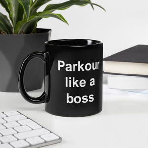 May include: Black ceramic mug with the text "Parkour like a boss" in white. The mug has a large handle and is sitting on a white surface. A plant and a book are in the background.