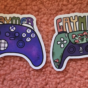 May include: Two stickers featuring game controllers with the word "GAYMER" above them. One controller is purple, the other is green with mushroom details. The stickers are on a textured, pink surface.