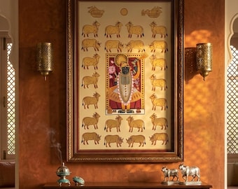 Puede incluir: Una obra de arte enmarcada que presenta una figura central rodeada de vacas doradas. La obra de arte se exhibe sobre un sofá con cojines estampados. Apliques decorativos de metal flanquean la obra de arte, y una mesa de madera contiene objetos decorativos.