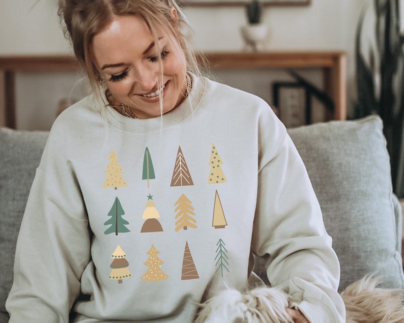 May include: A light beige sweatshirt with a pattern of stylized Christmas trees in shades of green, yellow, and brown.