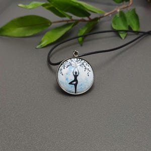 Può includere: Una collana con cordoncino nero con un ciondolo rotondo che presenta una silhouette nera di una persona che fa yoga con un albero sopra di lei. Il ciondolo ha uno sfondo bianco con accenti di acquerello blu.