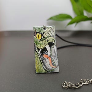 Op de afbeelding: Een rechthoekige hanger met een gedetailleerde illustratie van een open slangenbek met giftanden en een gespleten tong. De hanger is voornamelijk groen met gele en zwarte accenten en hangt aan een zwarte koordketting.