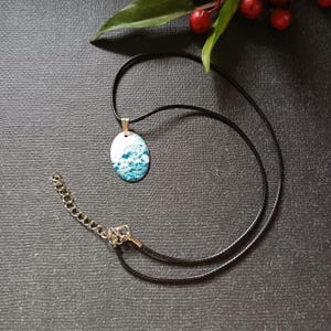 Peut inclure: Collier avec pendentif ovale représentant des vagues bleues et blanches. Le pendentif est suspendu à un cordon noir avec un fermoir et une chaîne d'extension dorés. Le collier est présenté sur une surface gris foncé, avec des baies rouges et des feuilles vertes en arrière-plan.