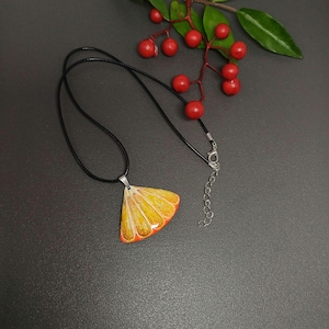 Può includere: Una collana con un cordoncino nero e una catena d'argento. Il ciondolo è una fetta d'arancia dipinta a mano con tonalità gialle e arancioni.