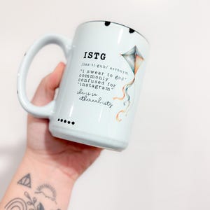 Pode incluir: Caneca de cerâmica branca com o acrónimo ISTG impresso, juntamente com a definição "I swear to god" e uma ilustração de pipa. A caneca tem uma alça grande e pontos pretos na borda.