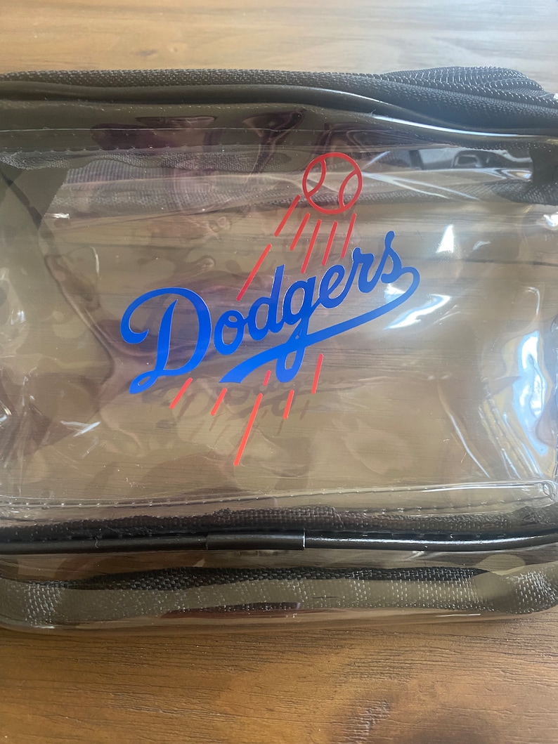 Pu&ograve; includere: Una borsa da viaggio rettangolare trasparente dei Dodgers con cerniera e finiture nere. Il logo dei Dodgers &egrave; in blu con un design a palla da baseball e strisce rosse. La borsa &egrave; in materiale trasparente.