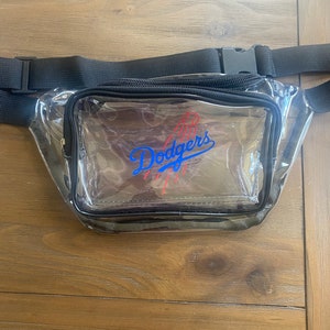 Pu&ograve; includere: Marsupio trasparente con cerniera nera e cinturino regolabile. La parte anteriore presenta il logo dei Los Angeles Dodgers in blu e rosso. Il marsupio &egrave; realizzato in plastica trasparente e ha un bordo nero.