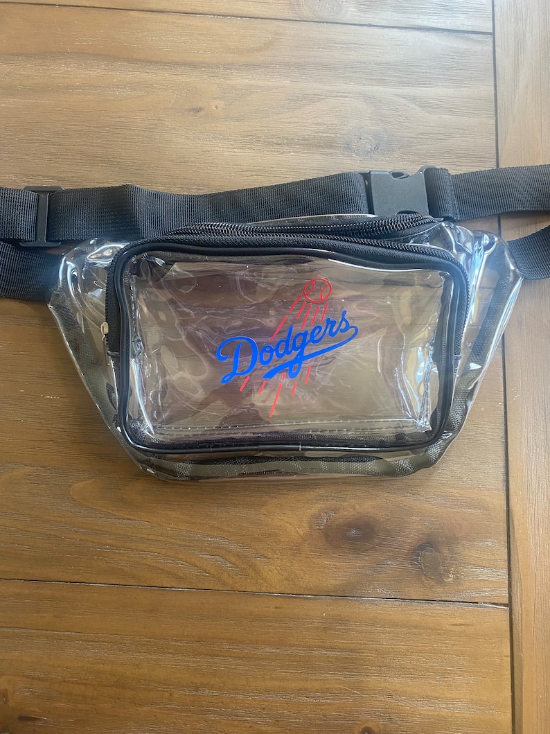 Pu&ograve; includere: Marsupio trasparente con cerniera nera e cinturino regolabile. La parte anteriore presenta il logo dei Los Angeles Dodgers in blu e rosso. Il marsupio &egrave; realizzato in plastica trasparente e ha un bordo nero.