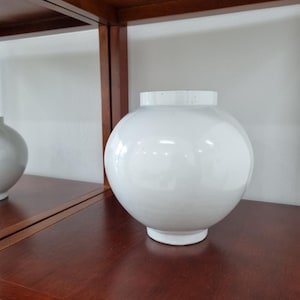 Puede incluir: Un jarrón esférico de cerámica blanca con un acabado liso y brillante y un cuello corto y cilíndrico. El jarrón presenta un cuerpo redondeado que se estrecha hacia una base circular pequeña, mostrando una superficie blanca sólida y limpia.