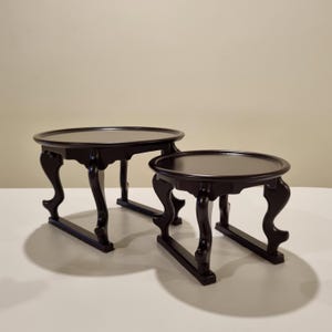 May include: Two dark brown wooden tables with smooth, circular tops and curved, ornate legs. Each table features a raised rim around the edge of the surface and a base structure where the legs connect to horizontal supports on the floor.