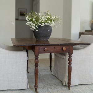 May include: A dark brown antique drop-leaf table with two drawers and turned legs. A black ceramic vase filled with white flowers sits on the table. Two upholstered chairs are on either side of the table.