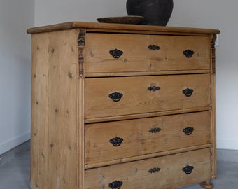 May include: A light-colored wooden dresser with four drawers and black hardware. The dresser has a decorative top and is topped with a dark, round pot and a small, round wooden plate. The dresser is made of wood and has a natural finish.