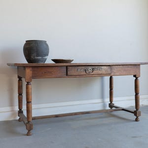 May include: A rectangular wooden table with a single drawer featuring an ornate metal pull and keyhole plate. The table has four turned legs connected by a lower stretcher. A dark gray rounded ceramic pot and a small shallow bowl sit on top.