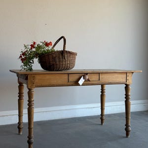 May include: A rectangular wooden table with four turned legs and a single central drawer featuring a round wooden knob. A woven brown wicker basket filled with green foliage and small red berries sits on the left side of the tabletop.