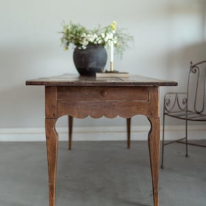 May include: A wooden antique table with a single drawer and scalloped apron. The table is topped with a dark vase of flowers and a candle. The table's legs are slender and curved. A metal chair is in the background.