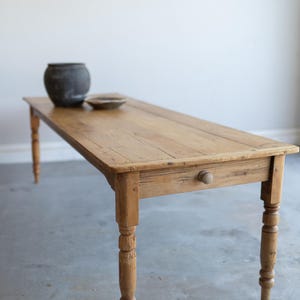 May include: A long, rectangular wooden table with a natural light brown finish, featuring a smooth tabletop, four turned legs, and a single drawer on the front side with a small, round wooden knob. A dark gray bowl sits on the table surface.