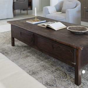 May include: A dark brown wooden coffee table with two drawers, an open book, a candle holder, and a decorative bowl. The table sits on a patterned rug in a living room setting. The table is approximately 48 inches long.