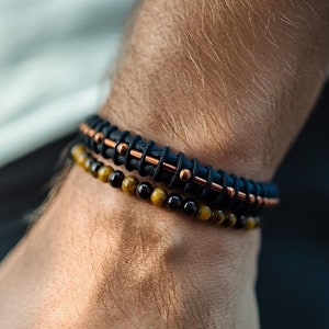 Könnte beinhalten: Zwei Armbänder an einem Handgelenk. Das obere Armband ist aus schwarzem Leder mit kupfernen Akzenten. Das untere Armband besteht aus Tigeraugenperlen.