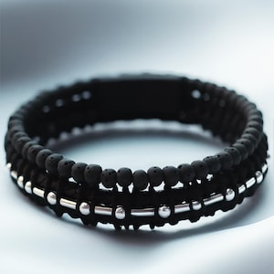 Op de afbeelding: Een gelaagde armband met zwarte lavastenen kralen en een zilveren band met zilveren kralen. De armband ligt op een wit oppervlak. "BLACK - SILVER - LAVA STONE" staat onder de armband.