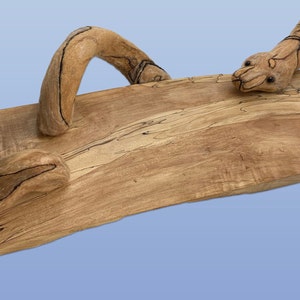 May include: A wooden serving tray featuring a carved snake design. The snake is curled around the edge of the tray and has a realistic appearance. The tray is crafted from natural wood with a light brown colour and a distinctive grain pattern.