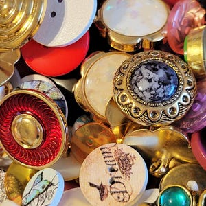 May include: A collection of assorted vintage buttons in various shapes, sizes, and colors. The buttons include gold, red, white, and iridescent designs. Some buttons have floral and butterfly motifs, while others feature intricate patterns.
