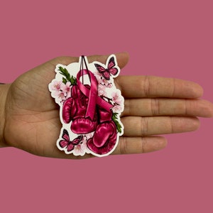 May include: Pink boxing gloves with a pink ribbon and flowers. The gloves are tied together with the ribbon. There are two butterflies on the sticker.