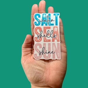 May include: A rectangular sticker with the words "Salt Water Sea Shells Shine" in blue, coral, and pink. The sticker is held in a hand against a teal background. The words are stacked vertically.