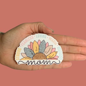 May include: A white sticker with a colorful sunflower design. The sunflower has pink, yellow, and blue petals. The words "strong, fearless, loving, warm, selfless" are written around the sunflower. The word "mom" is written in cursive at the bottom of the sticker.