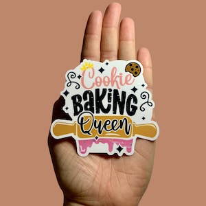 May include: White sticker with a brown rolling pin, pink icing, and black text that reads "Cookie Baking Queen" with a gold crown and a brown cookie.
