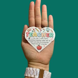 May include: A heart-shaped sticker with the word "TEACHER" in colorful letters and a definition of the word. The sticker is decorated with colorful school-themed icons, including an apple, a globe, a pencil, and a ruler.