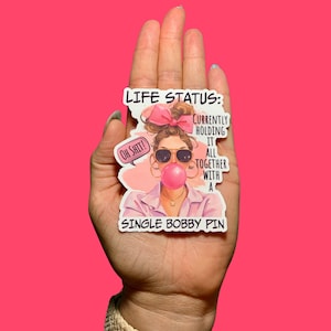 May include: A sticker with a cartoon woman with pink hair and sunglasses blowing a bubble. The text on the sticker reads "LIFE STATUS: CURRENTLY HOLDING IT ALL TOGETHER WITH A SINGLE BOBBY PIN".