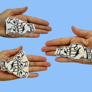 May include: Black and white stickers featuring a fist bump design. The stickers depict a large fist bumping two smaller fists, with the word "DAD" on the wrist. The stickers are on a light blue background.