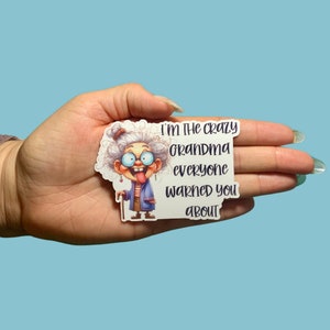May include: A sticker featuring a cartoon image of a gray-haired grandma with glasses and her tongue sticking out. The text on the sticker reads, "I'M THE CRAZY GRANDMA EVERYONE WARNED YOU ABOUT."