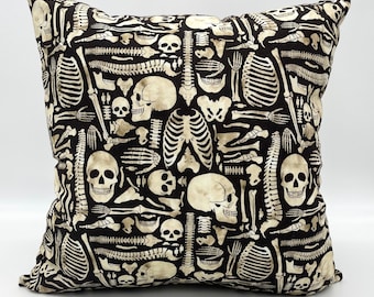 May include: Square decorative pillow with a black background and a repeating pattern of off-white human skeletal elements, including skulls, ribs, and vertebrae. The pillow is likely made of fabric and is designed for home decor.