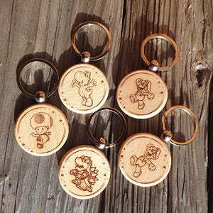 Könnte beinhalten: Eine Sammlung von sechs Holzschlüsselanhängern mit gravierten Cartoon-Charakteren. Jeder Schlüsselanhänger hat einen silberfarbenen Ring und einen runden Holzanhänger mit einem anderen Charakterdesign. Die Schlüsselanhänger werden auf einer verwitterten Holzoberfläche präsentiert.