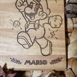 Puede incluir: Una placa de madera con una imagen grabada con l&aacute;ser de Mario, un personaje popular de videojuegos, en una pose de carrera. La placa presenta el texto "MARIO" y elementos decorativos como estrellas y un hongo.