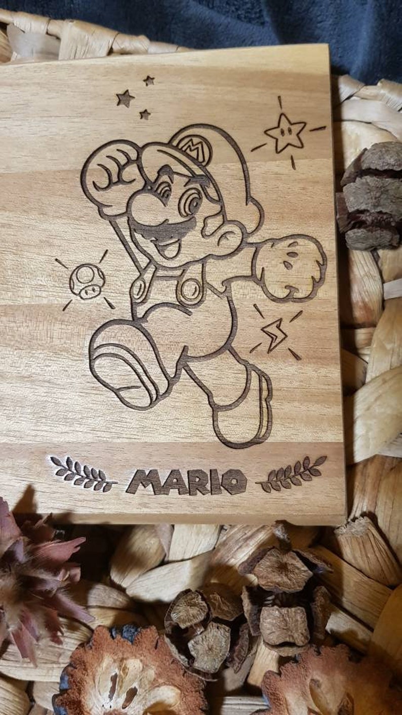 Puede incluir: Una placa de madera con una imagen grabada con l&aacute;ser de Mario, un personaje popular de videojuegos, en una pose de carrera. La placa presenta el texto "MARIO" y elementos decorativos como estrellas y un hongo.
