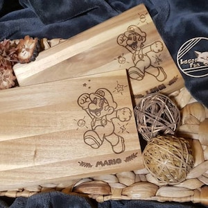Puede incluir: Dos tablas de cortar de madera con una imagen grabada con l&aacute;ser de Mario del videojuego Super Mario Bros. Las tablas est&aacute;n sobre una superficie de mimbre tejido.