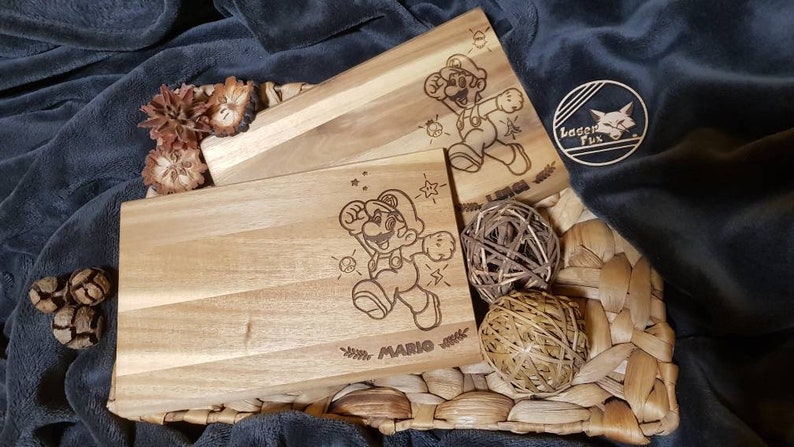 Puede incluir: Dos tablas de cortar de madera con una imagen grabada con l&aacute;ser de Mario del videojuego Super Mario Bros. Las tablas est&aacute;n sobre una superficie de mimbre tejido.