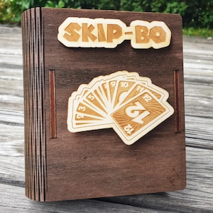 Könnte beinhalten: Dunkelbraune Skip-Bo-Kartenspielbox aus Holz mit dem Spielnamen in hellbrauner Schrift. Die Box hat einen Fächer aus Holzkarten. Die Box besteht aus geschichtetem Holz.
