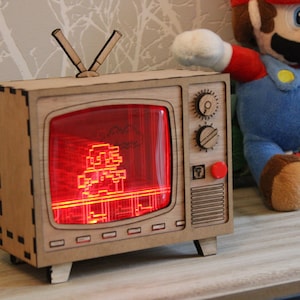 May include: A wooden retro-style television with a red screen displaying a pixelated image of a video game character. The TV has knobs, a red button, and an antenna. A plush Mario doll is in the background.