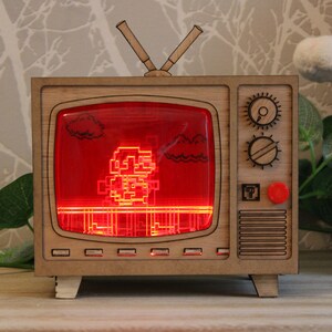 May include: A wooden TV-shaped lamp with a red screen displaying a pixelated character. The lamp has a retro design with knobs and antennas. The character is a pixelated figure, likely from a video game. The lamp is a decorative item.