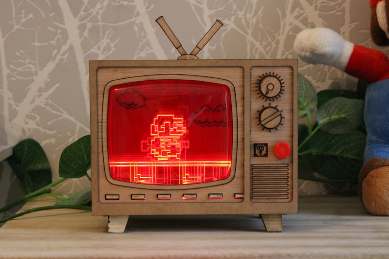 May include: A wooden TV-shaped lamp with a red screen displaying a pixelated character. The lamp has a retro design with knobs and antennas. The character is a pixelated figure, likely from a video game. The lamp is a decorative item.