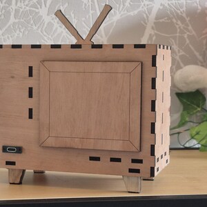 May include: A wooden, retro-style television set with a rectangular frame and antenna. The TV has a light brown finish with a darker brown frame around the screen. The design includes small, rectangular cutouts and is set on four small legs.