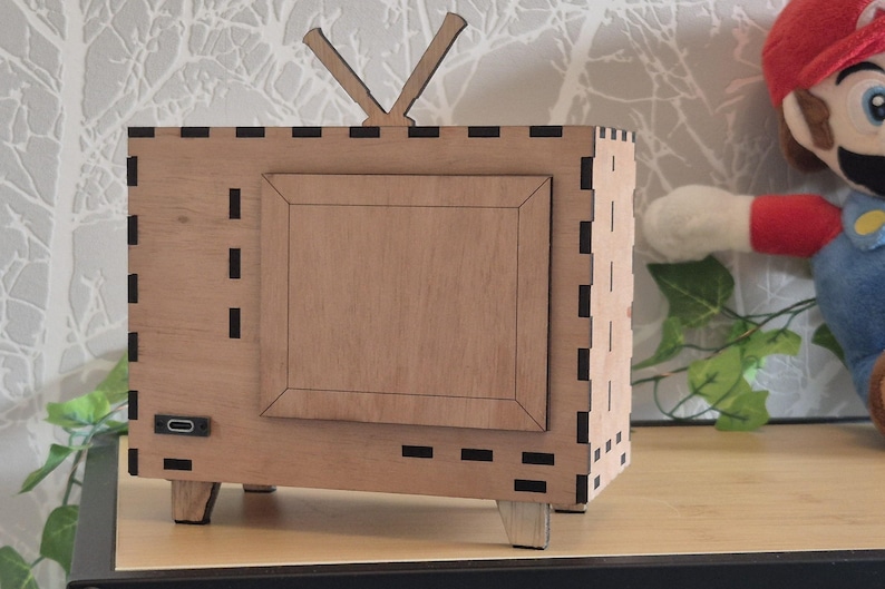 May include: A wooden, retro-style television set with a rectangular frame and antenna. The TV has a light brown finish with a darker brown frame around the screen. The design includes small, rectangular cutouts and is set on four small legs.