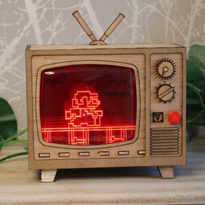 May include: Wooden TV-shaped night light featuring a pixelated video game character on a red screen. The light has a retro design with antenna and control knobs. A red button and a small square emblem are on the side.
