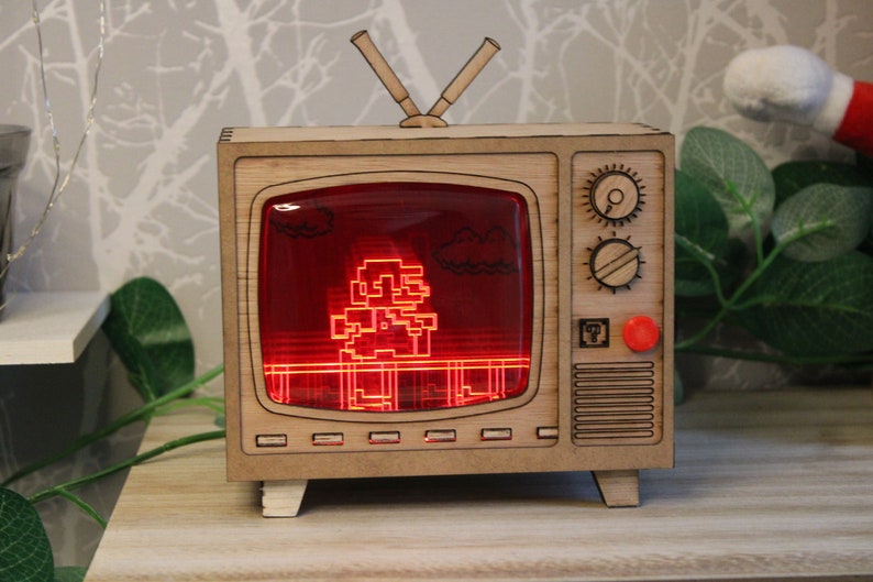 May include: Wooden TV-shaped night light featuring a pixelated video game character on a red screen. The light has a retro design with antenna and control knobs. A red button and a small square emblem are on the side.